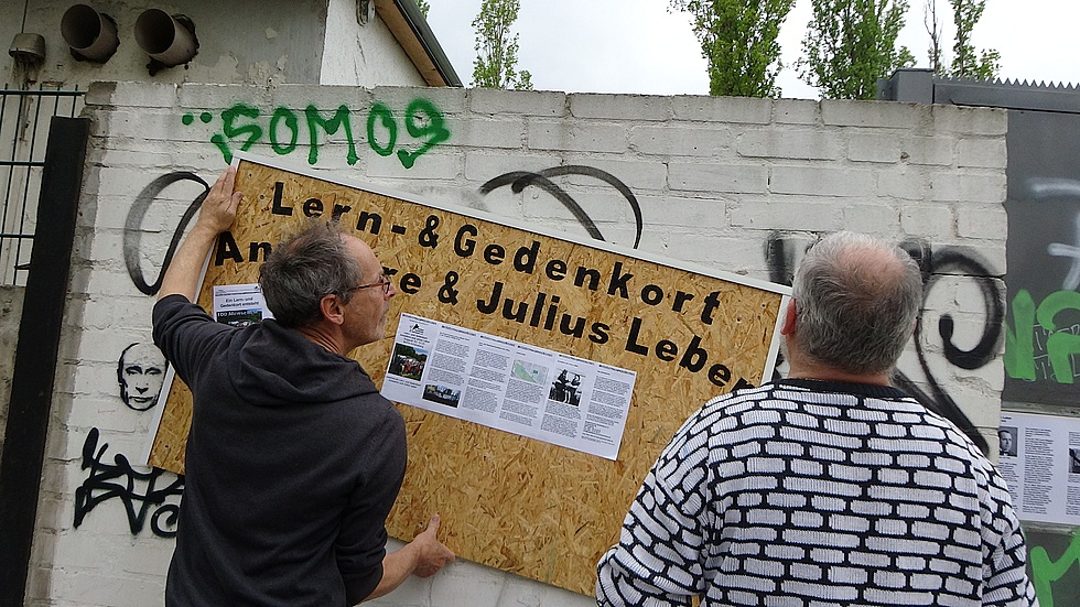 Zwei Mitglieder des Arbeitskreises hängen eine Tafel an der ehemaligen Kohlenhandlung auf