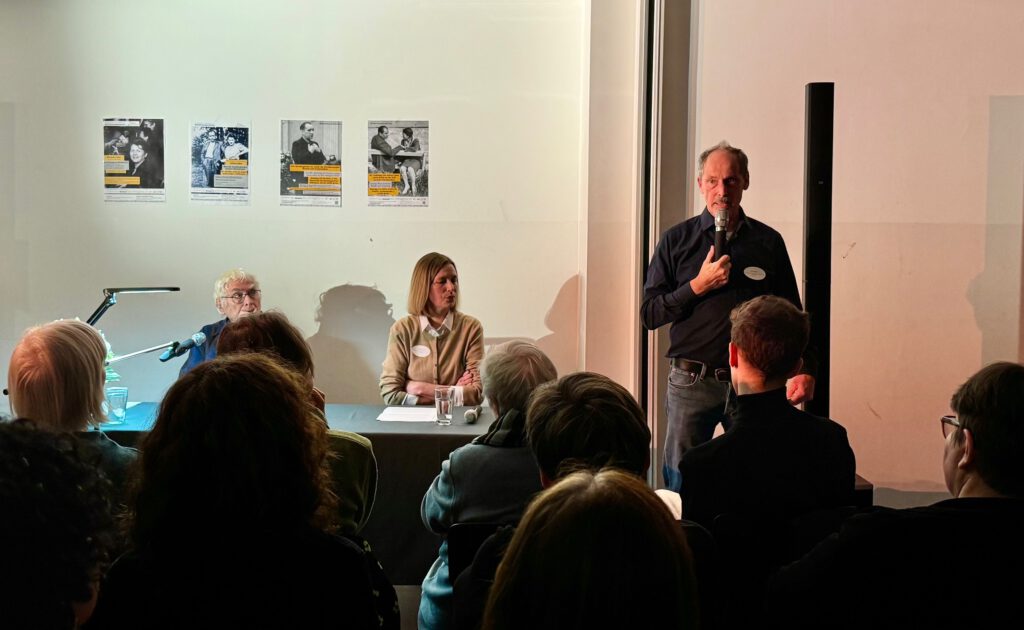 Zwei Menschen am Tisch, einer stehend mit Mikrofon vor Publikum