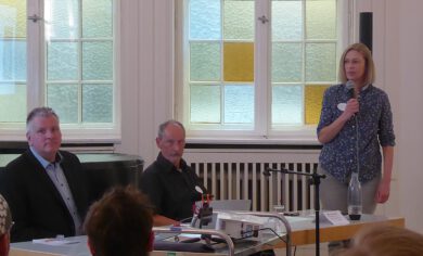 Frau mit Mikrofon und zwei Männer am Podium